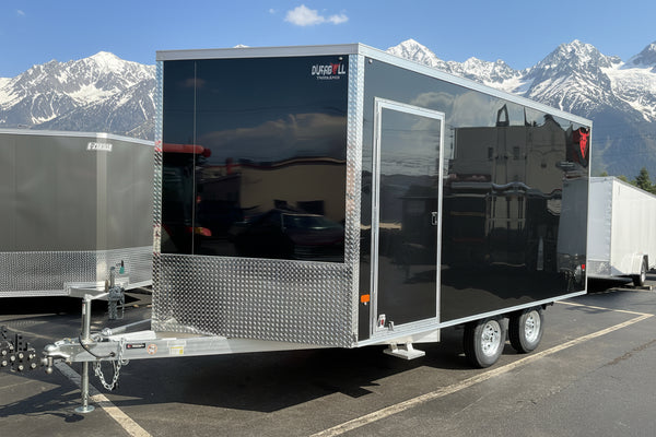 A black enclosed cargo trailer with silver aluminum sides, diamond plate bumpers, and a side entry door, parked outdoors in a lot with other trailers and a building in the background.