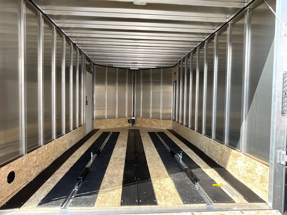 A black enclosed cargo trailer with silver aluminum sides, diamond plate bumpers, and a side entry door, parked outdoors in a lot with other trailers and a building in the background.