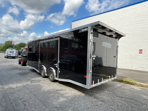 A black enclosed car trailer with silver aluminum framing and side windows, parked outdoors on asphalt beside a white building under a blue sky with clouds.