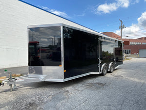 A black enclosed cargo trailer with silver aluminum bumper and hitch hardware, parked outdoors on a paved lot next to a white brick building under a blue sky.