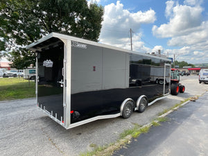 A black and silver enclosed race trailer parked outdoors on a paved lot under a blue sky with clouds. The rear door is open, showing the interior, and a tractor is visible behind the trailer.