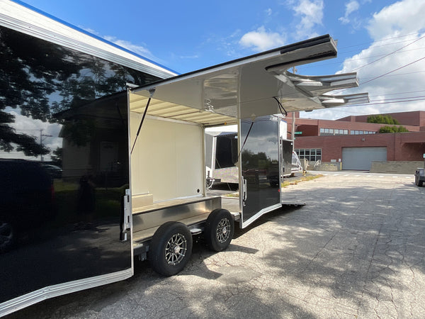 A black and stainless steel mobile food truck trailer parked outdoors on a paved lot under a blue sky. The side compartment is open, showing a cream-colored interior, with a metal awning extended above.
