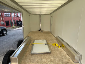 The inside of a large cargo trailer or shipping container, showing beige walls, a wood-look floor, metal side rails, and a closed white door in the center. A yellow strap is tied across the floor, and a red brick building and part of a black vehicle are visible outside through the open side door.