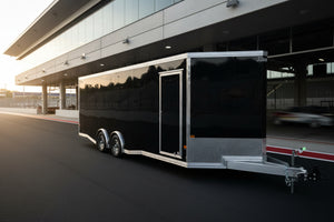A black and silver enclosed car trailer parked outdoors on a paved lot, with trees, buildings, and a dark pickup truck visible in the background.