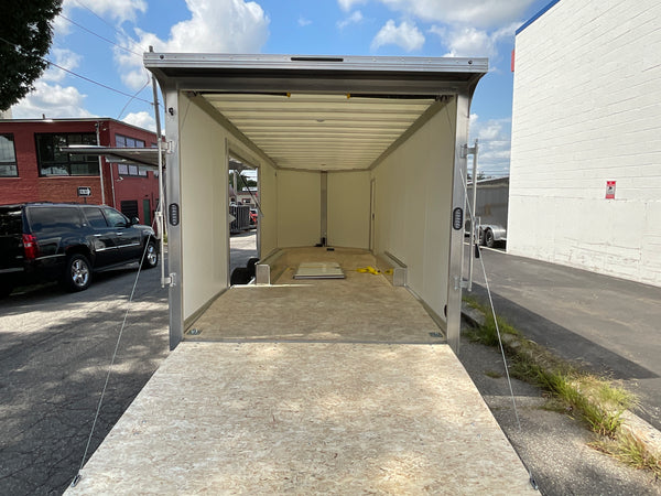 An empty enclosed cargo trailer with its rear ramp open, photographed outdoors between buildings. The trailer has a silver metal frame, cream-colored interior walls, and a plywood floor.