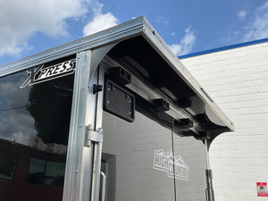 Close-up of the rear entry area of a silver and black enclosed trailer, showing the Xpress model name, a black license plate frame, and the High Country logo against a white brick building and blue sky.