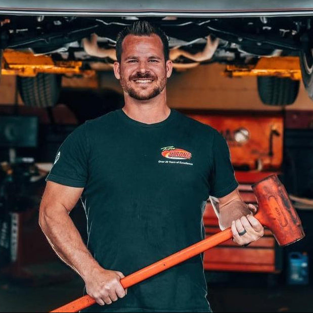 Man holding a tool under a car in a garage with a 'Toy Shop' sign in the background.