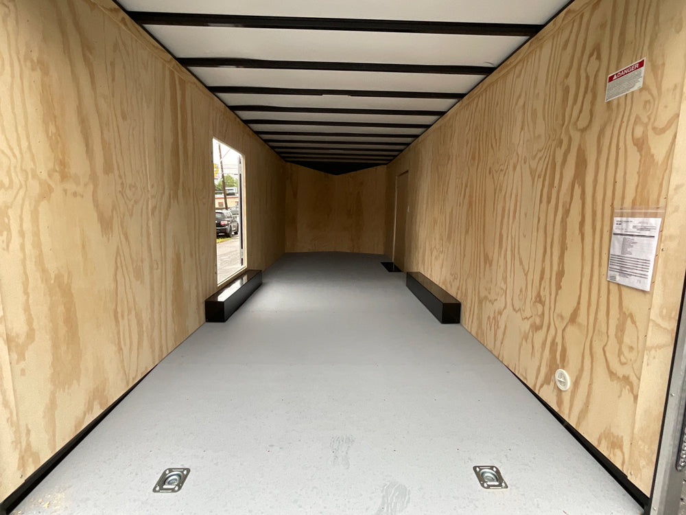 A black enclosed cargo trailer with silver trim, a side door, and a diamond-plate lower front bumper, parked outdoors on wet asphalt next to a red brick building.