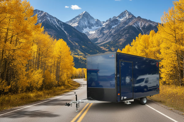 A dark blue enclosed cargo trailer with a diamond-plate lower section and a side entry door, parked outdoors on a paved lot with a red trailer and warehouse building in the background.