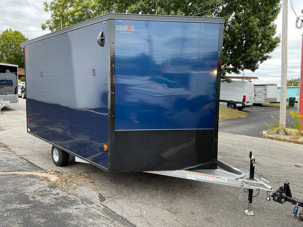 A dark blue enclosed trailer parked outdoors on a paved lot, with a black diamond-plate lower section, silver metal hitch, and visible reflections on the side door.