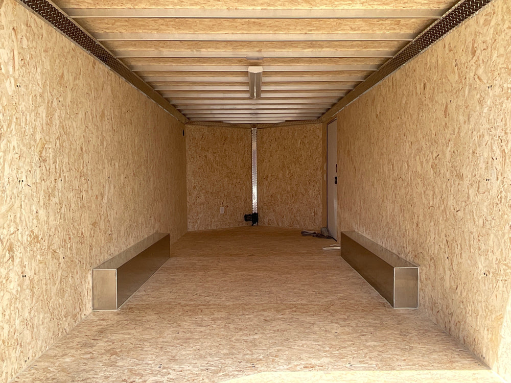 A black enclosed cargo trailer with silver aluminum framing, a rear ladder, and a side door, parked outdoors on a paved surface with trees and a blue sky in the background.