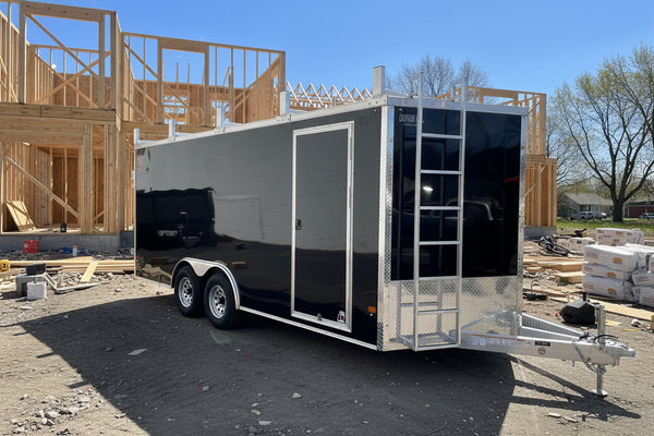A black enclosed cargo trailer with silver aluminum framing, a rear ladder, and a side door, parked outdoors on a paved surface with trees and a blue sky in the background.