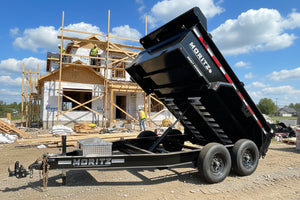 A black Moritz dump trailer with its dump bed raised, parked outdoors in a dealership lot with other trailers and a building in the background.