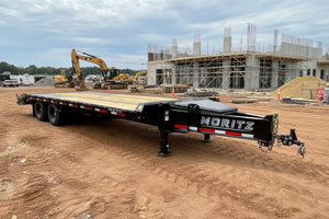 A black Moritz flatbed trailer with wooden decks and a diamond plate front, parked on an outdoor lot with other trailers and buildings in the background.