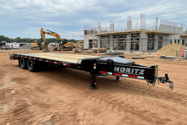 A black Moritz flatbed trailer with wooden decks and a diamond plate front, parked on an outdoor lot with other trailers and buildings in the background.