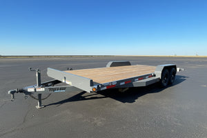 A low-profile tandem flatbed trailer with a wood deck and gray metal frame, parked on asphalt in an outdoor dealership lot.