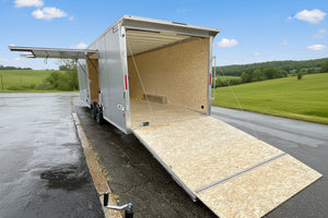 A large silver enclosed cargo trailer with its rear ramp lowered, showing a plywood-lined interior, parked outdoors on wet pavement between buildings. A white Colliers van and another vehicle are visible in the background.