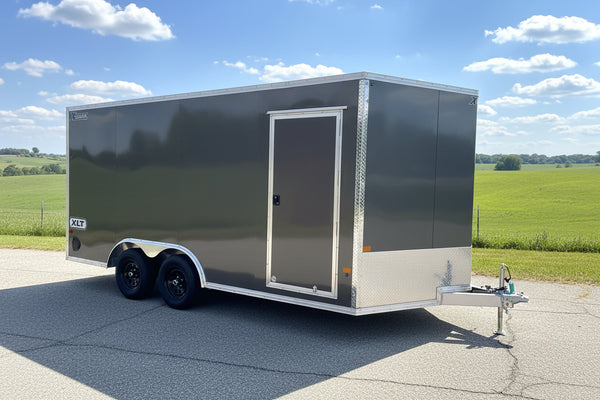 A charcoal aluminum enclosed cargo trailer parked outdoors on a paved road. The trailer has a side door with a silver frame, dual wheels, a hitch at the front, and silver diamond-plate metal trim at the corners and along the lower side.