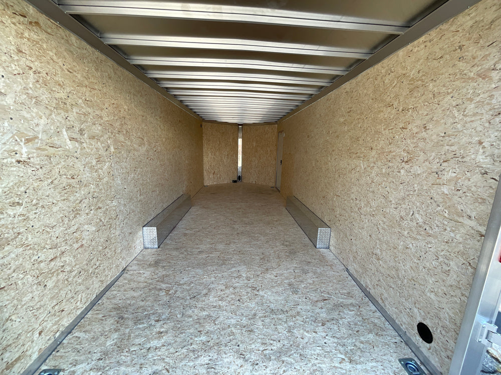 A white enclosed cargo trailer parked outdoors on a paved lot next to a red building. The trailer has a side door, a diamond-plate lower section, dual wheels, and a hitch at the front.